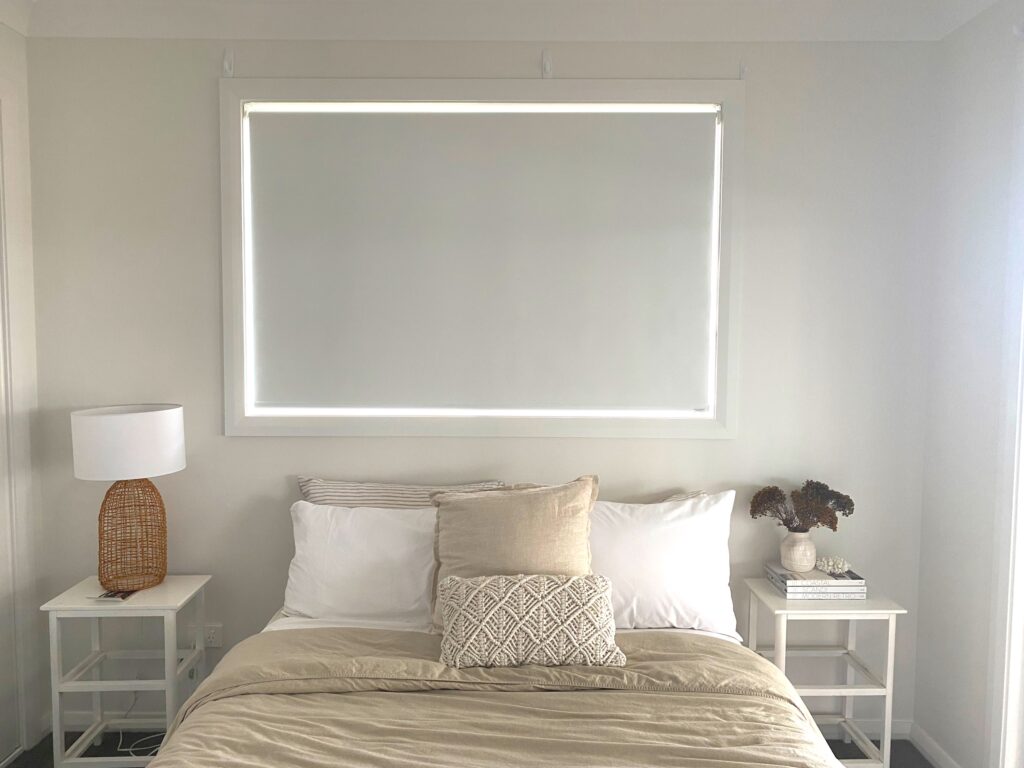 A minimalist bedroom featuring a bed with neutral-toned linens, flanked by two white nightstands, positioned directly under a window with a closed white roller blind.