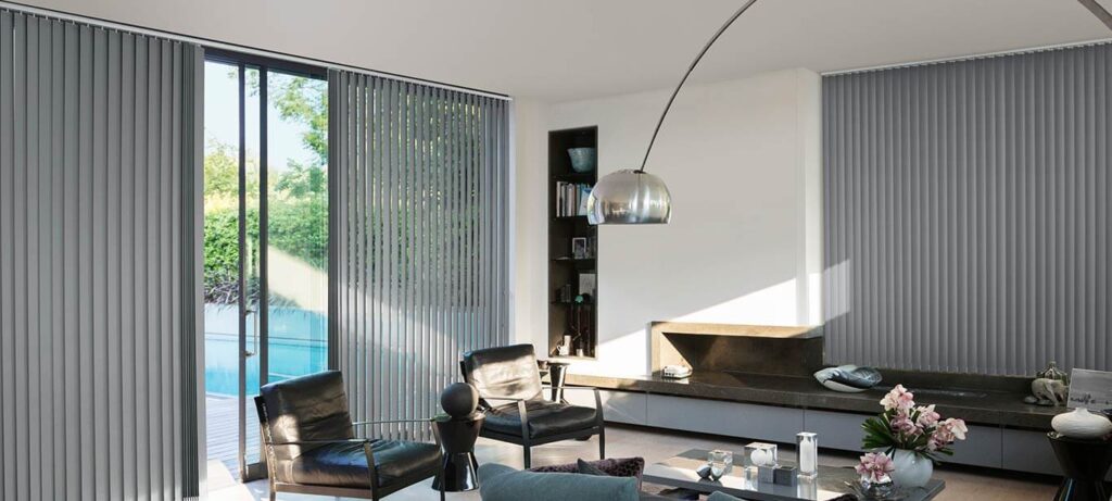 A modern, stylish living room featuring black leather armchairs, an arched chrome floor lamp, and grey vertical blinds framing large glass doors that open to a pool area.