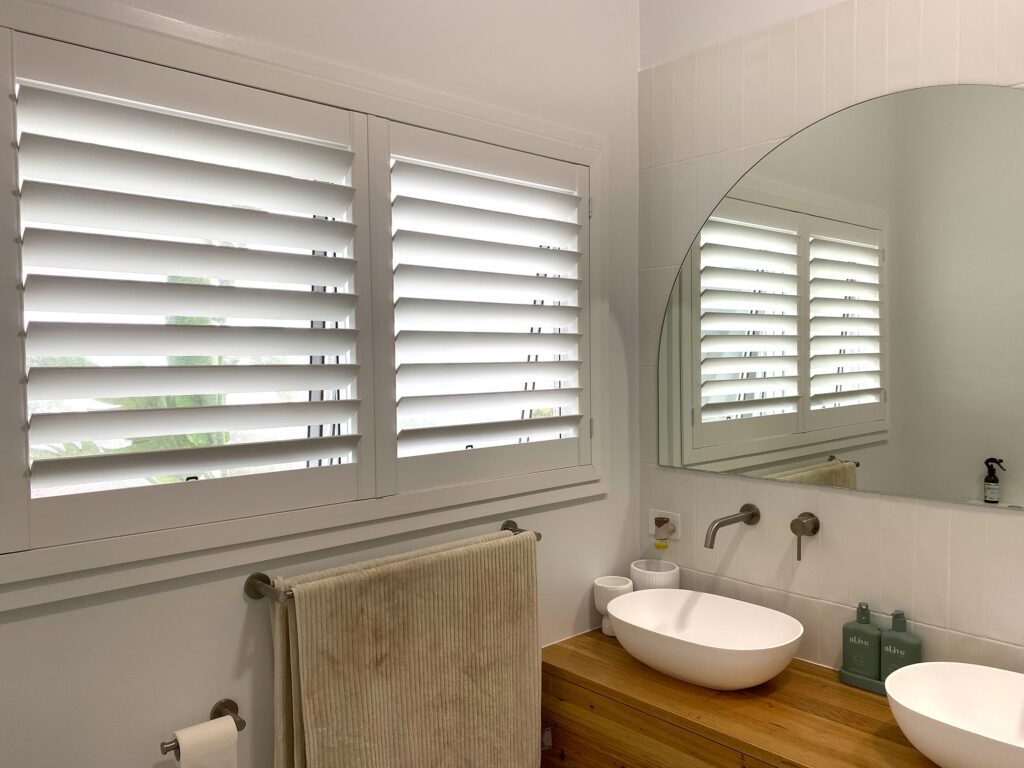A modern bathroom vanity featuring a wooden countertop with two vessel sinks, a large arched mirror, and a window fitted with white plantation shutters.