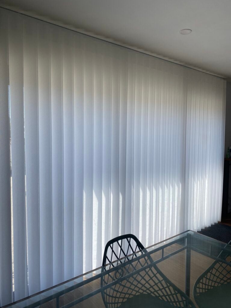 A dining room with a glass-top table, featuring a large window expanse entirely covered by floor-to-ceiling sheer white vertical blinds that filter incoming sunlight.