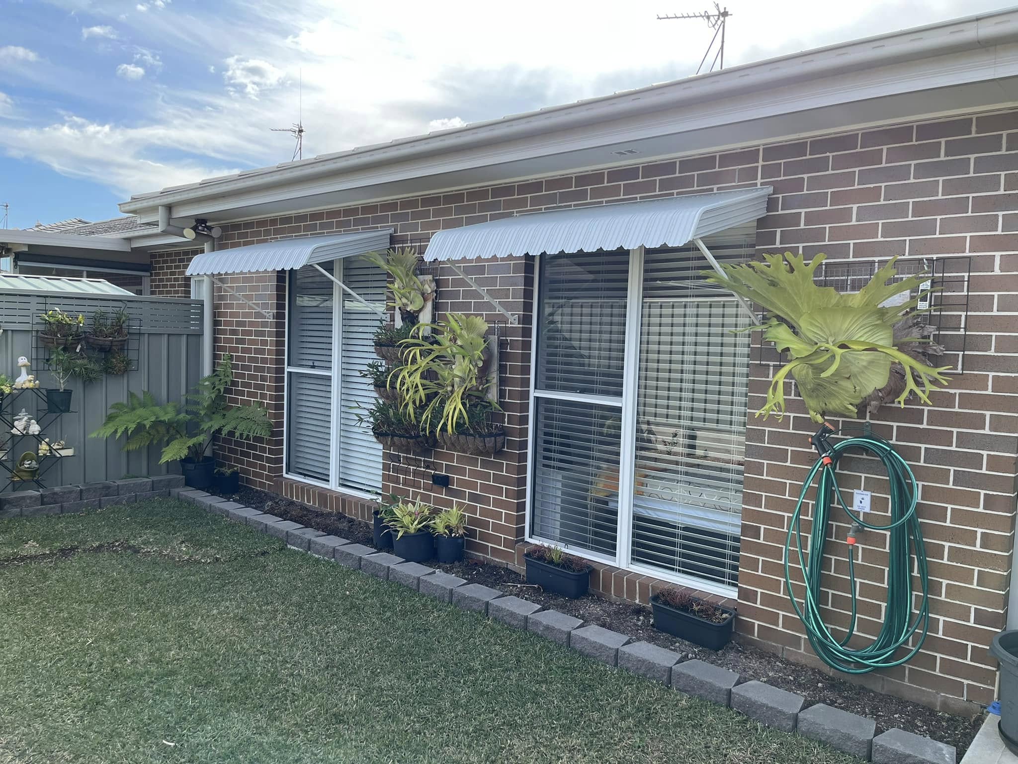 The backyard exterior of a brick house featuring white metal window awnings over two large windows with blinds.