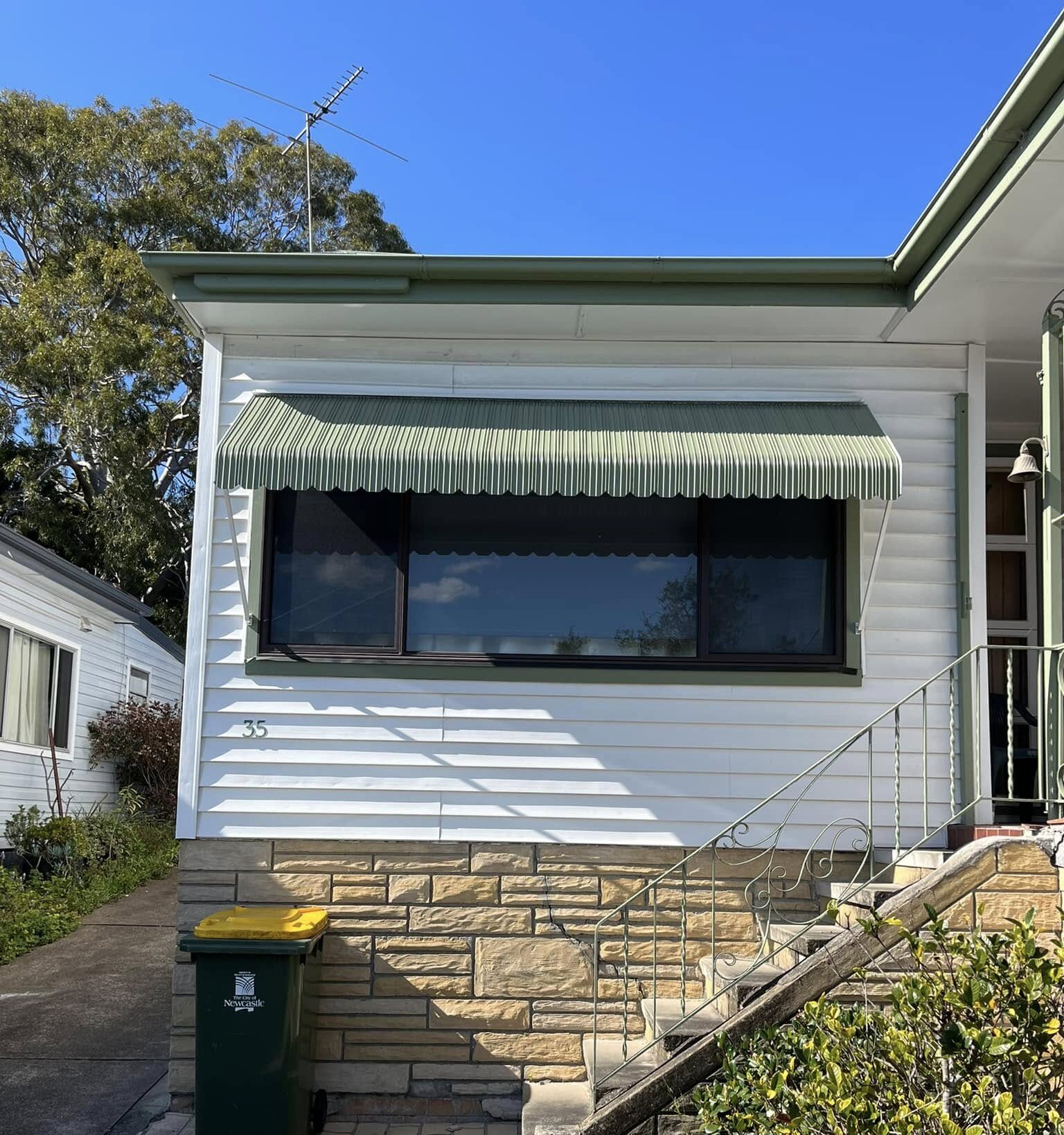 An exterior view of a white weatherboard house with a stone foundation, featuring a wide green metal window awning with a scalloped edge installed over a large front window next to concrete entry steps.