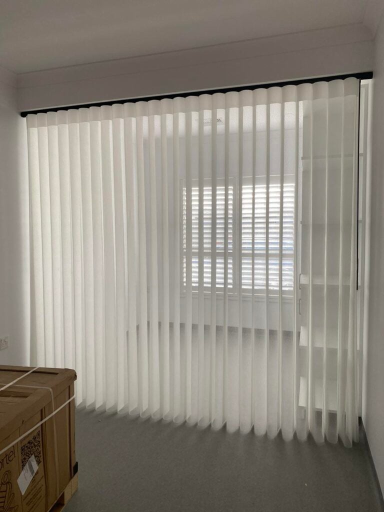 A room showing white sheer vertical smart privacy blinds hanging from a black ceiling track, partially veiling a window with white plantation shutters and a storage shelf.
