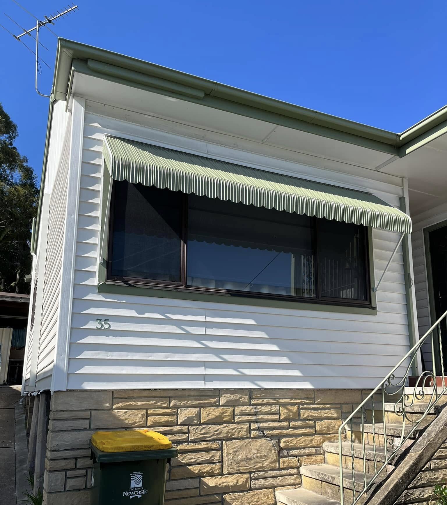 An angled exterior view of a white weatherboard house showing a green metal window awning with a scalloped trim mounted over a large window.
