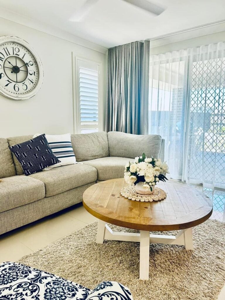 A living room featuring a light grey sectional sofa, a round wooden coffee table with white flowers, a large wall clock, and sliding doors dressed with sheer white curtains and grey drapery.