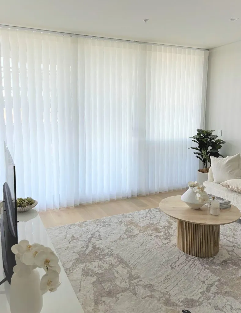 A bright, minimalist living room featuring a light-wood fluted coffee table on a patterned area rug, with a large glass door covered by floor-to-ceiling sheer white vertical drapes.