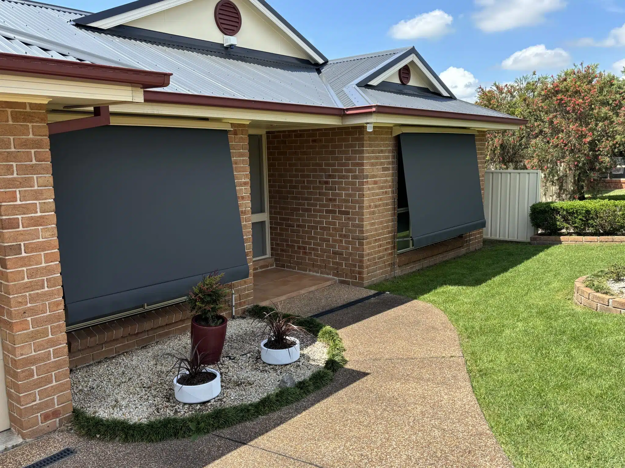 An exterior front view of a brick house with a concrete walkway and green lawn, featuring two dark charcoal canvas automatic lock-arm awnings.