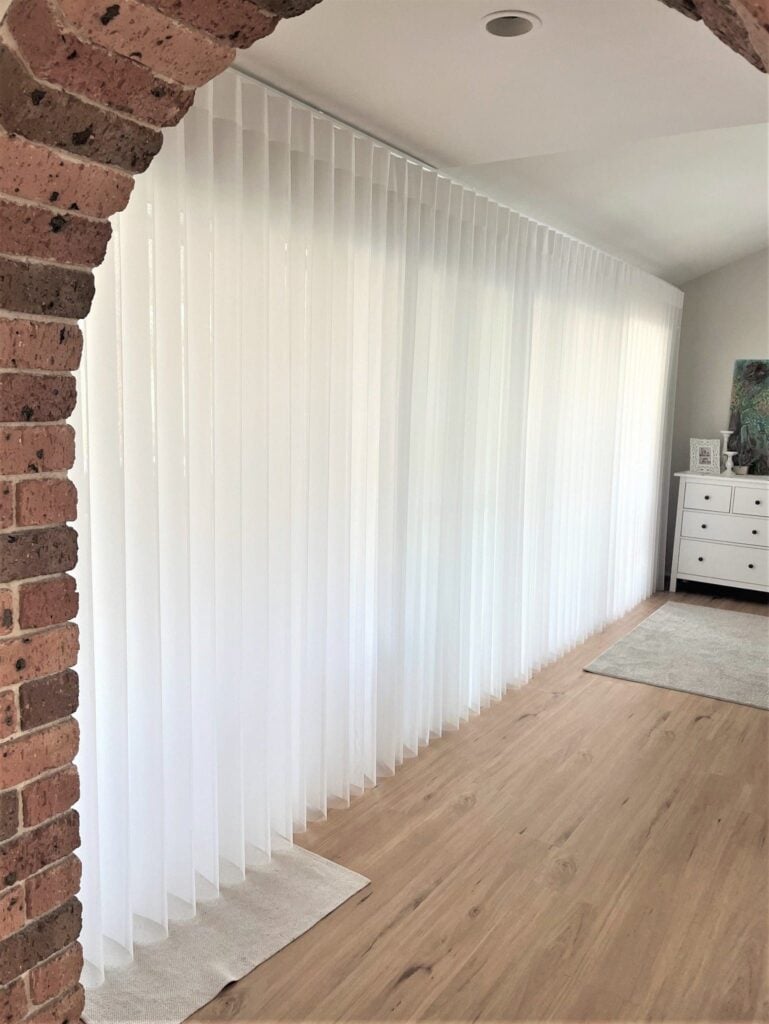 An indoor hallway view framed by a brick archway, showing light wood floors and a very long wall covered by white vertical sheer blinds.