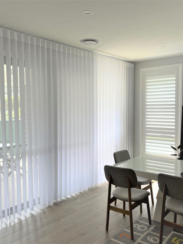 A bright dining space featuring a white table and grey upholstered chairs next to a large sliding glass door covered by floor-to-ceiling sheer white vertical blinds.