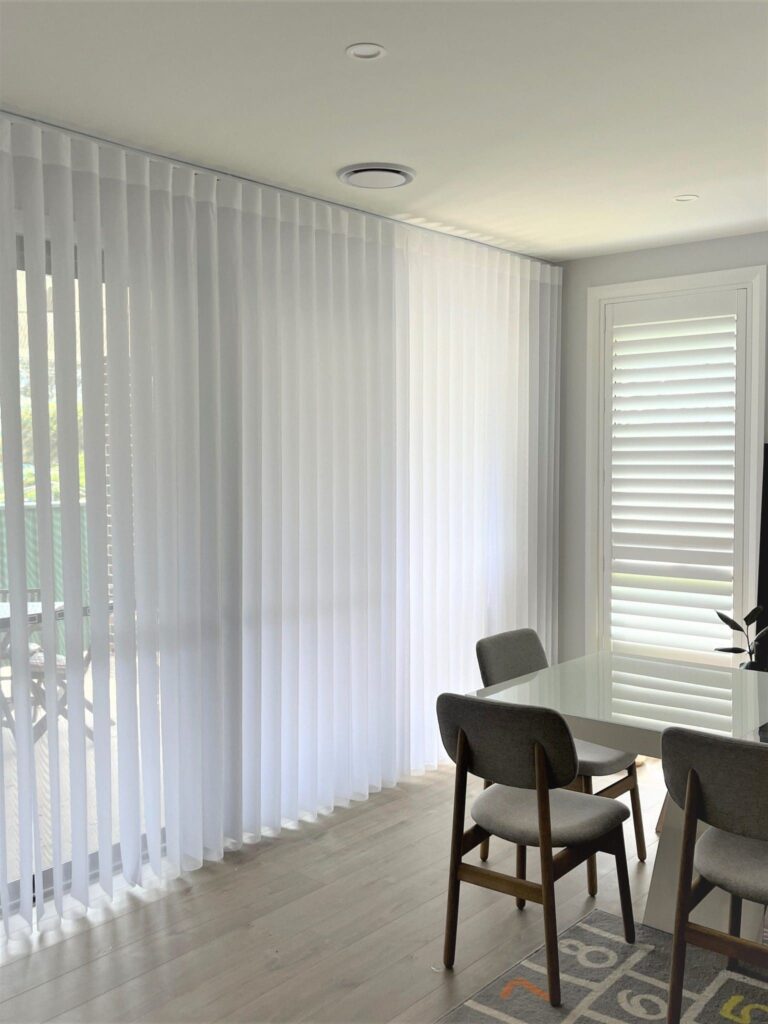A bright dining room featuring a white table with grey upholstered chairs, a white plantation shutter window, and large sliding glass doors covered by white vertical sheer blinds.