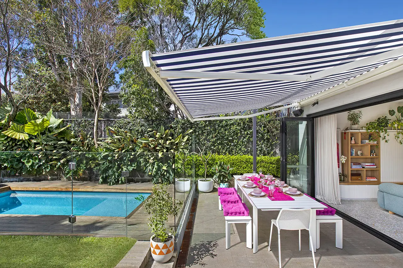 A vibrant patio dining area set up next to a swimming pool with a glass fence, featuring a navy-and-white striped retractable awning.