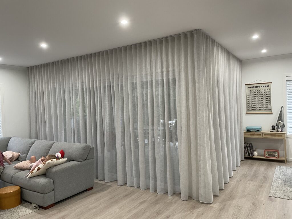 A living room with a grey sectional sofa, showcasing a large corner track setup with floor-to-ceiling light grey sheer S-fold curtains wrapping around a glass door area.