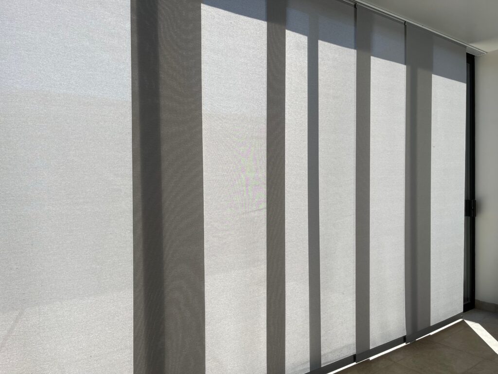 A wide glass sliding door covered by light grey vertical panel glide blinds, casting shadows on the tiled floor from the bright daylight outside.