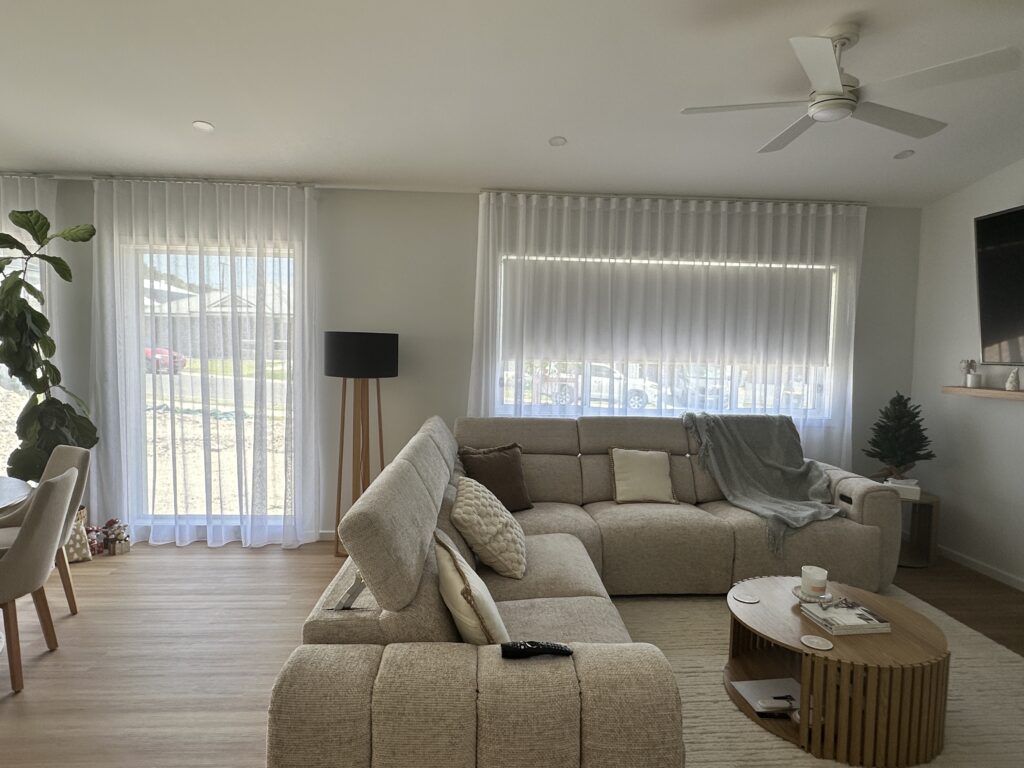 A bright, modern living room with a large beige sectional sofa, a wooden coffee table, and a ceiling fan, featuring a glass door and window dressed with light, sheer white S-fold curtains.