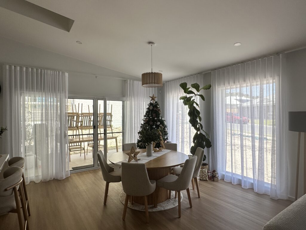 A bright dining room featuring a round wooden table and multiple large windows and glass doors framed by long, sheer white S-fold curtains.