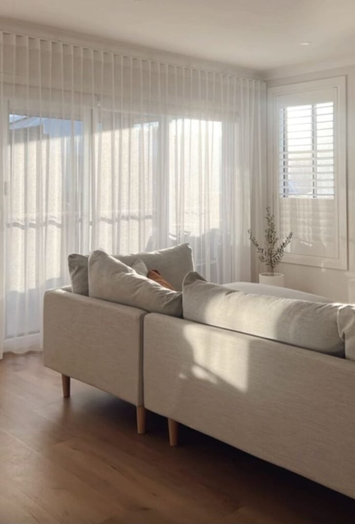 A sunlit living room with a large sliding glass door covered by floor-to-ceiling sheer white wave-fold curtains and a side window fitted with white plantation shutters.