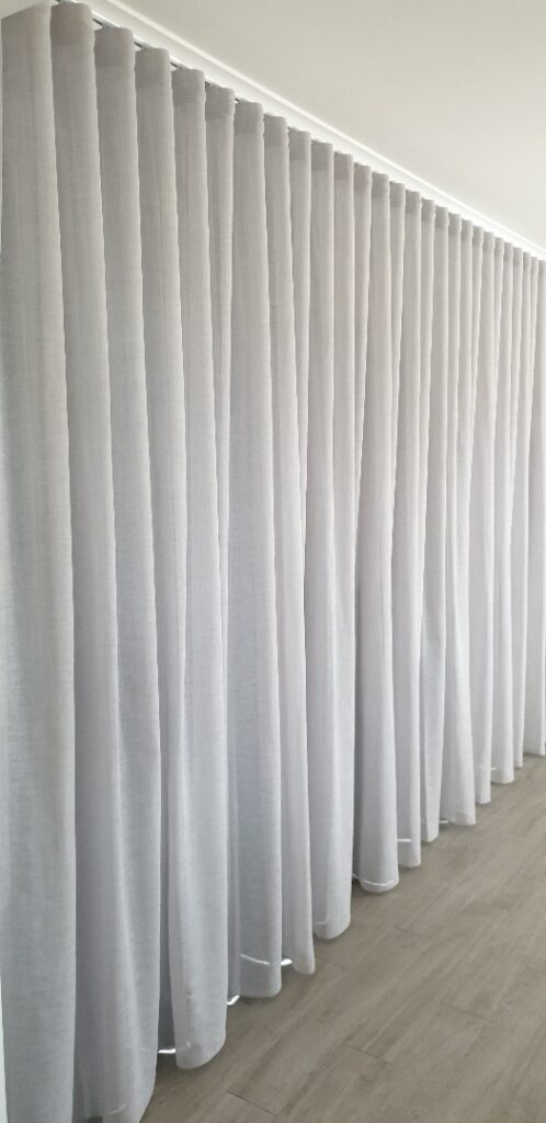 A full-length view of a long wall draped with light grey, textured S-fold sheer curtains hanging from a sleek ceiling track down to a wood-look floor.