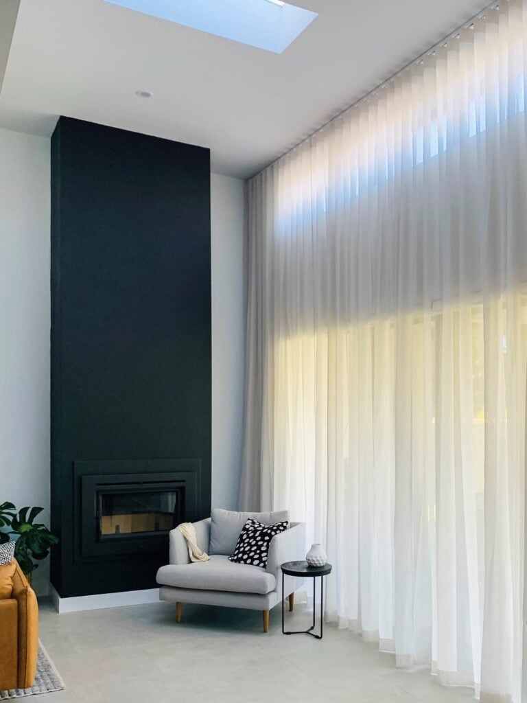 A modern, high-ceiling living room featuring a tall black fireplace structure, a light grey armchair, and a massive wall of floor-to-ceiling sheer white curtains filtering the light.
