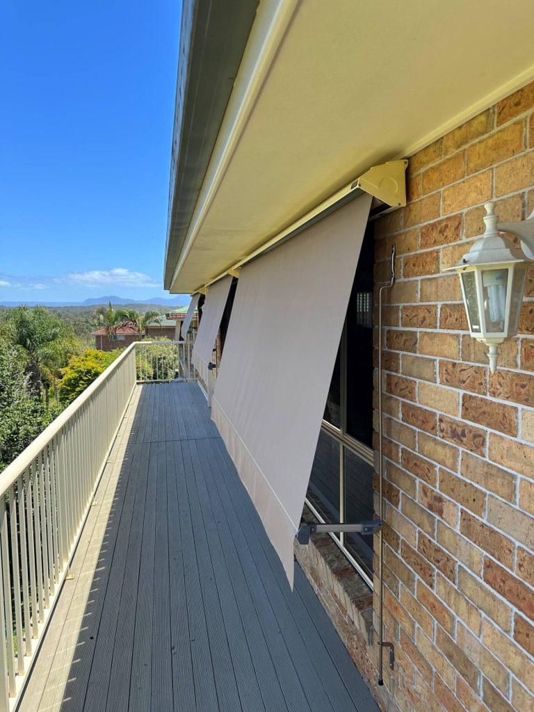 A long elevated wooden balcony with beige railings on a brick building, featuring a series of tan automatic lock-arm awnings.