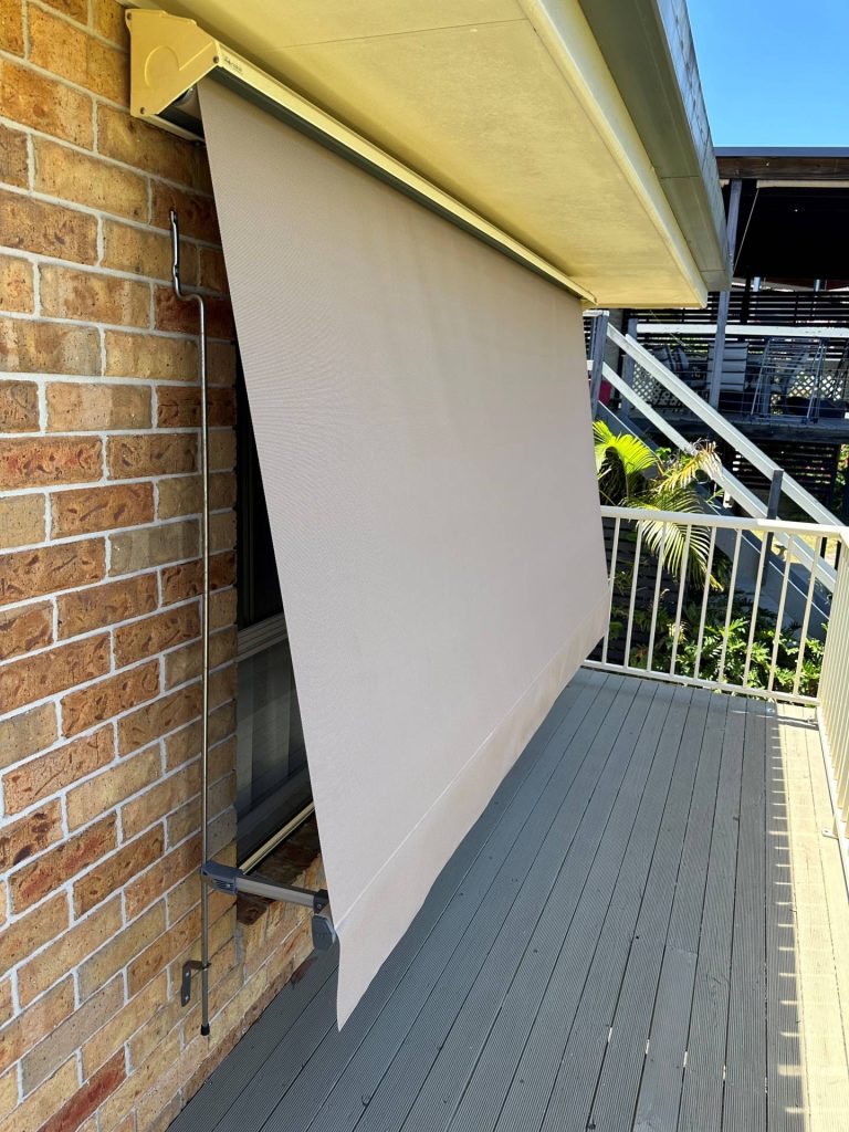 A tan automatic lock-arm awning lowered over a window along a wooden balcony deck on a brick building.