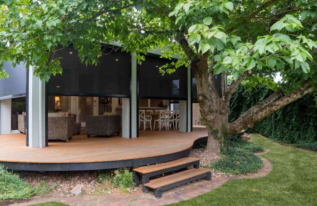 A curved, elevated wooden deck shaded by a large tree, featuring outdoor wicker seating and a dining set enclosed by dark mesh privacy blinds.