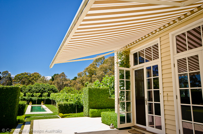 A sunlit patio outside a yellow weatherboard house featuring a large tan-and-white striped retractable awning.