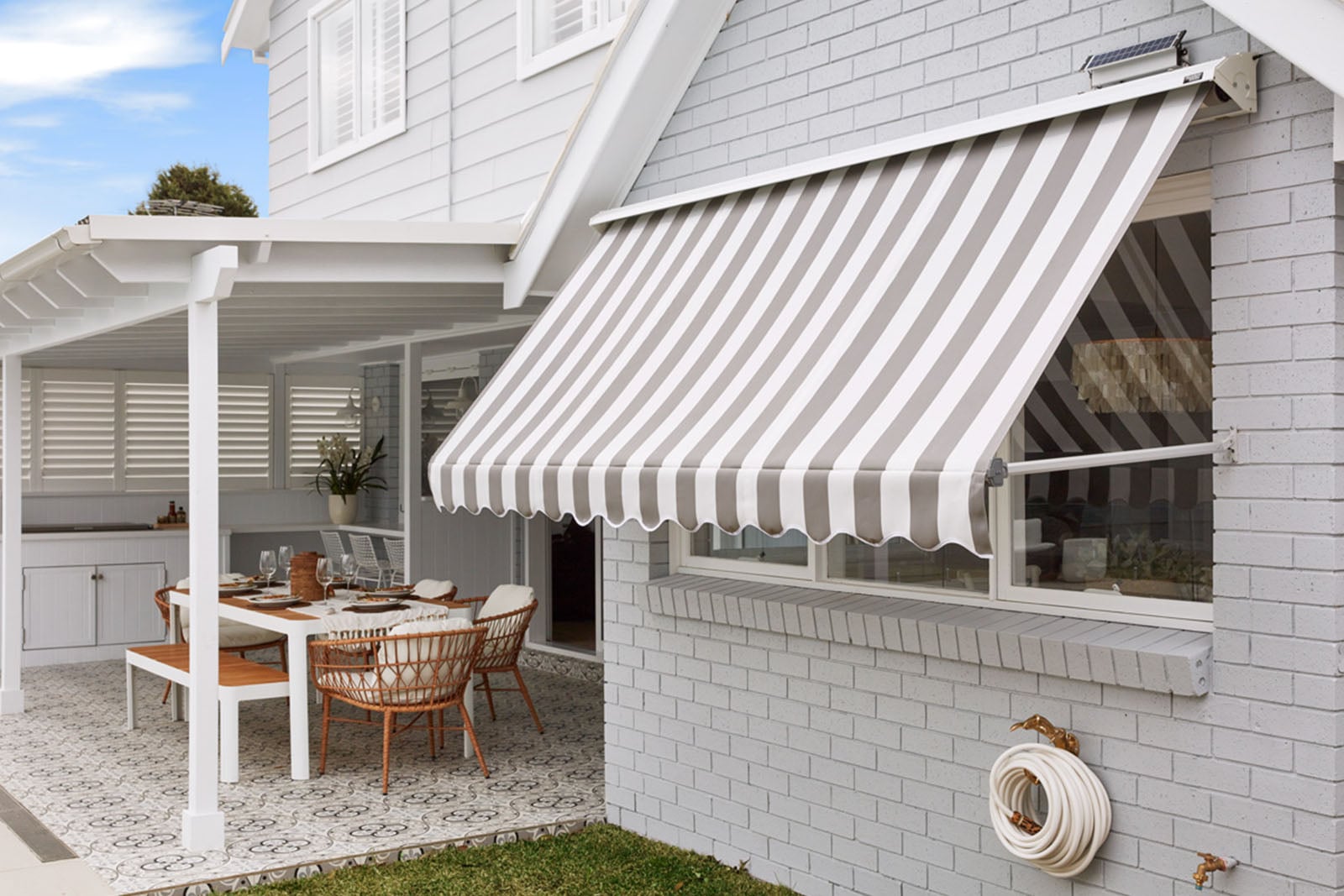 An exterior view of a light grey brick house, featuring a grey and white striped retractable awning extended over a window next to a covered patio with an outdoor dining table.