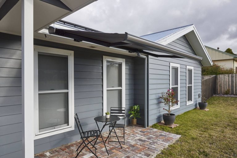 A small brick-paved patio outside a grey weatherboard house featuring a black folding-arm retractable awning.