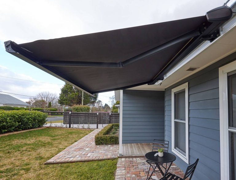 A large black retractable folding-arm awning fully extended over a brick patio with a small bistro set, attached to a grey siding house with a backyard lawn.