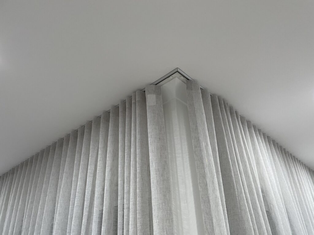A close-up, low-angle shot of a white ceiling meeting a corner, showing light grey S-fold sheer curtains mounted onto a sleek corner ceiling track.