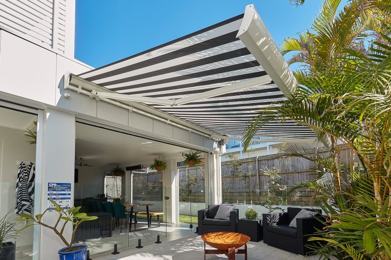 A sunlit, modern outdoor patio and lounge area shaded by a large black-and-white striped retractable awning.