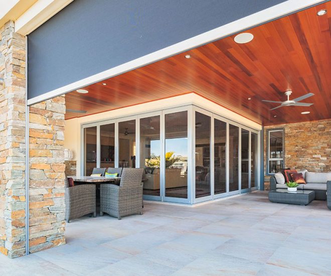 A spacious outdoor patio with a rich wooden ceiling, stone pillar and walls, outdoor seating, and a large grey motorised mesh shade partially lowered over the opening.