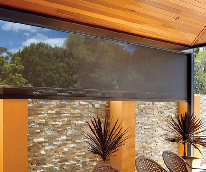An outdoor dining patio under a polished timber ceiling with a fan, featuring wicker chairs at a tiled table and a dark mesh privacy shade partially lowered in front of a stone accent wall.
