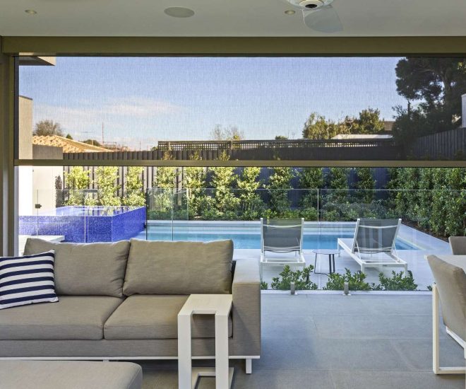 A covered modern patio with grey sofa seating and a dining set, featuring a dark mesh outdoor shade partially lowered to frame a view of a backyard swimming pool and lounge chairs.
