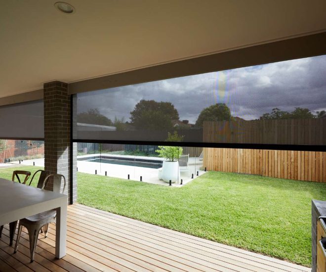 A covered wooden deck with a dining table and outdoor kitchen, featuring dark motorised mesh privacy shades partially lowered over a view of a grassy backyard and swimming pool.