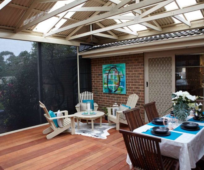 A covered wooden deck featuring an exposed timber truss ceiling, a set dining table, and an outdoor seating area with Adirondack chairs enclosed by a dark privacy mesh screen.