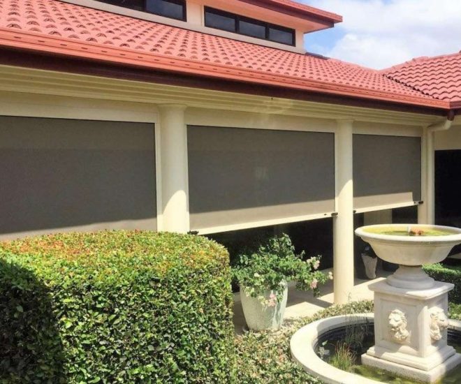An exterior view of a building with a red tiled roof, manicured garden hedges, and a lion-themed fountain, showing dark outdoor roller blinds pulled down between cream-coloured pillars.