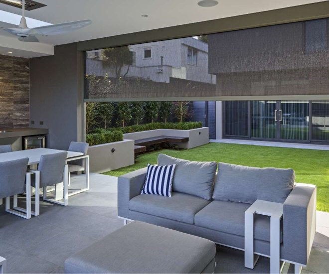 A covered modern patio with a grey outdoor sofa, dining set, and built-in kitchenette, featuring a dark mesh privacy shade partially lowered above a manicured backyard lawn.
