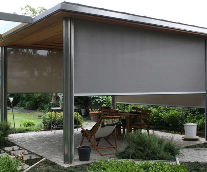 A freestanding garden gazebo with metal pillars and a paved floor, featuring an outdoor dining set and lounge chair partially enclosed by large, grey roller privacy shades.