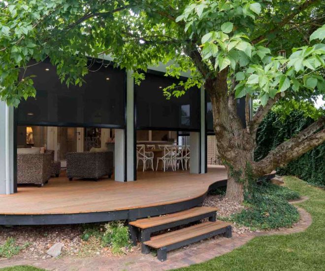A curved, elevated wooden deck shaded by a large tree, featuring outdoor wicker seating and a dining set enclosed by dark mesh privacy blinds.