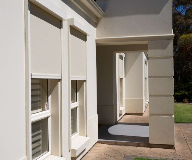 An exterior view of a cream-coloured house with a tiled walkway, showcasing windows equipped with colour-matched outdoor roller blinds partially drawn to block sunlight.