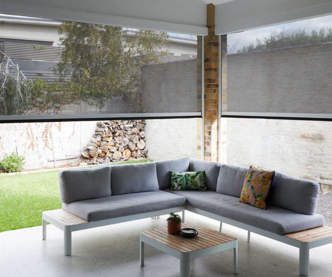 A covered patio featuring a grey sectional sofa with decorative pillows and a wooden coffee table, enclosed by large motorised mesh privacy shades looking out onto a small grassy yard.