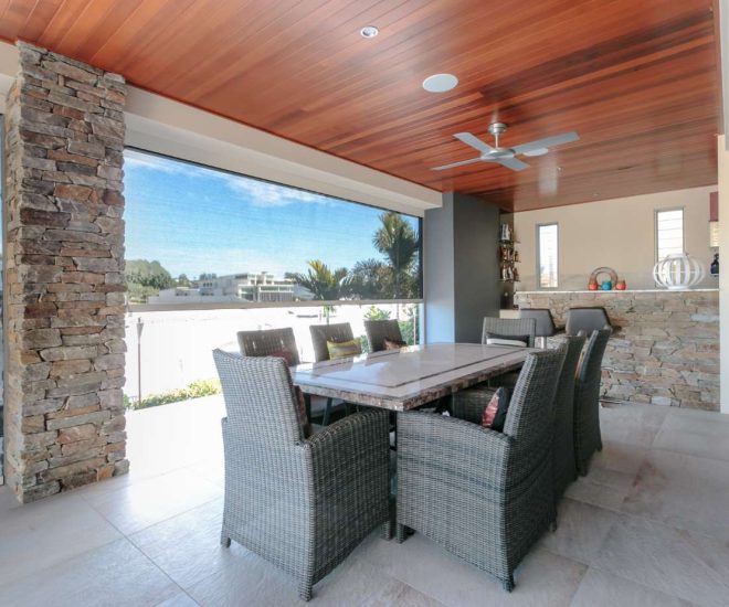 A luxurious outdoor patio featuring a polished timber ceiling with light-coloured motorised mesh shades partially lowered to overlook a waterfront view.