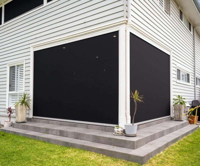 An exterior view of a two-storey house with light grey siding, showing a corner patio fully enclosed by large, solid black outdoor roller blinds.