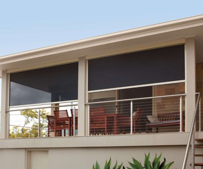 An elevated outdoor balcony patio with a stainless steel cable railing, wooden dining and lounge furniture, and dark outdoor privacy shades partially lowered under a white ceiling.