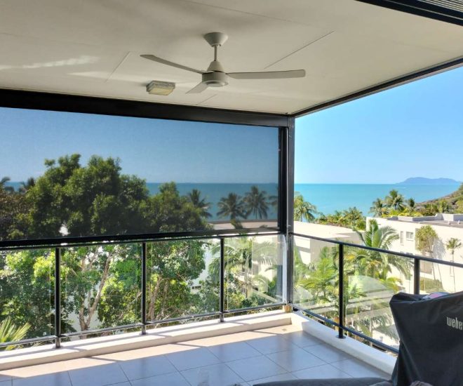 A covered apartment balcony with a white ceiling fan and a covered Weber grill, featuring a dark mesh outdoor shade partially lowered to filter sunlight while preserving an expansive ocean view.