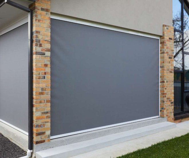 An exterior view of a modern house with brick pillars, showing large, solid grey outdoor roller blinds fully lowered to enclose a patio area.