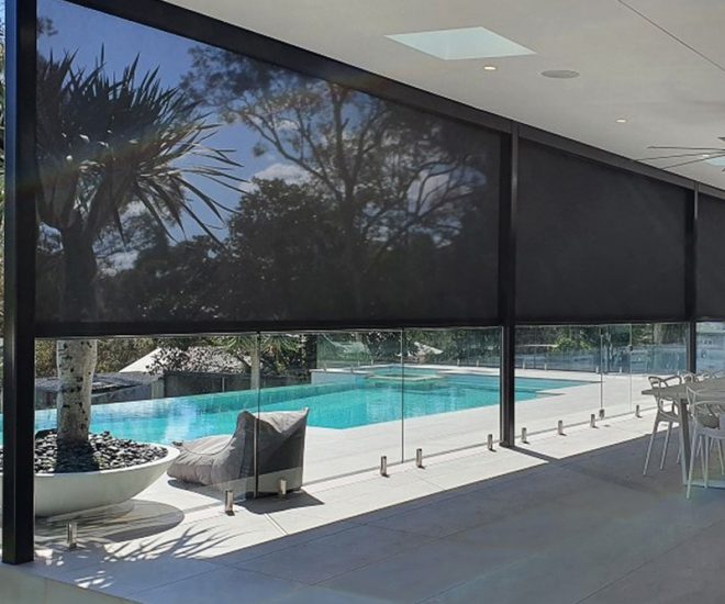 A luxury outdoor patio with black motorised mesh privacy shades partially lowered, overlooking a glass-fenced swimming pool area.