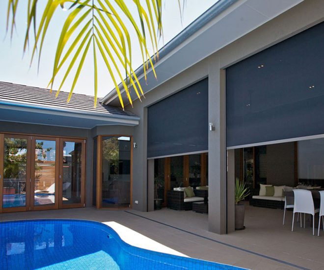 A backyard swimming pool alongside a covered outdoor living area with a patio dining set, lounge chairs, and large, dark outdoor privacy shades partially lowered.